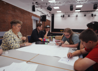 Eine pädagogische Fachkraft sitzt an einem Tisch mit Jugendlichen bei einem Workshop. Die Jugendlichen beschriften Plakate. 