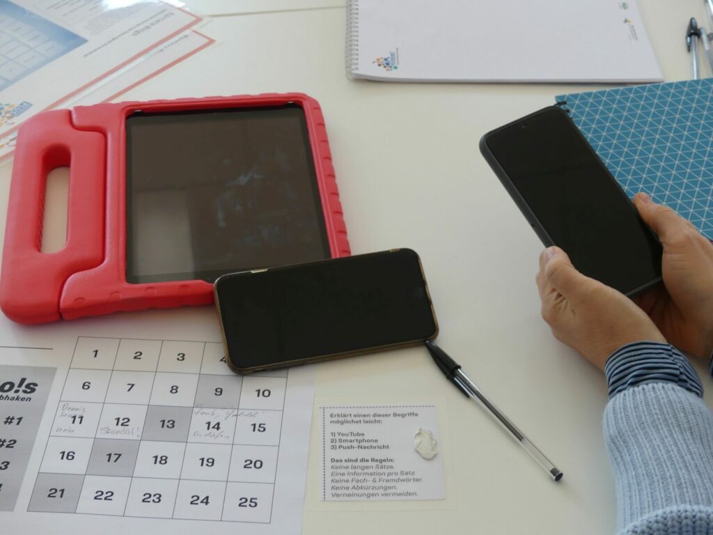 Auf einem Tisch liegen ein Tablet und ein Smartphone. Außerdem sieht man den ausgedruckten Bogen vom Barriere-Bingo. Zwei Hände halten ein weiteres Smartphone. 